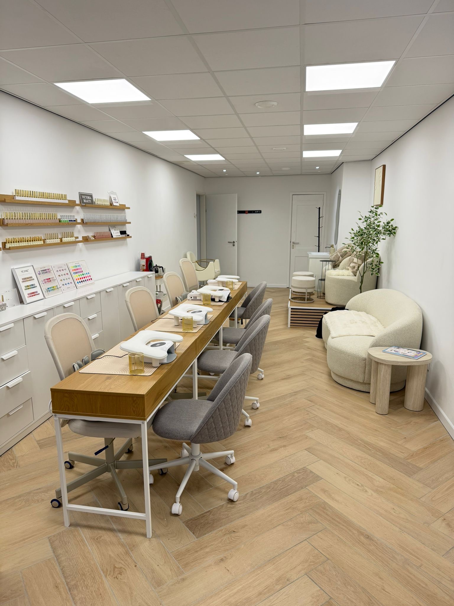 De studio van Victoria beauty: een lange houten werktafel met meerdere werkplekken, visgraatvloer en een zithoek bij het raam